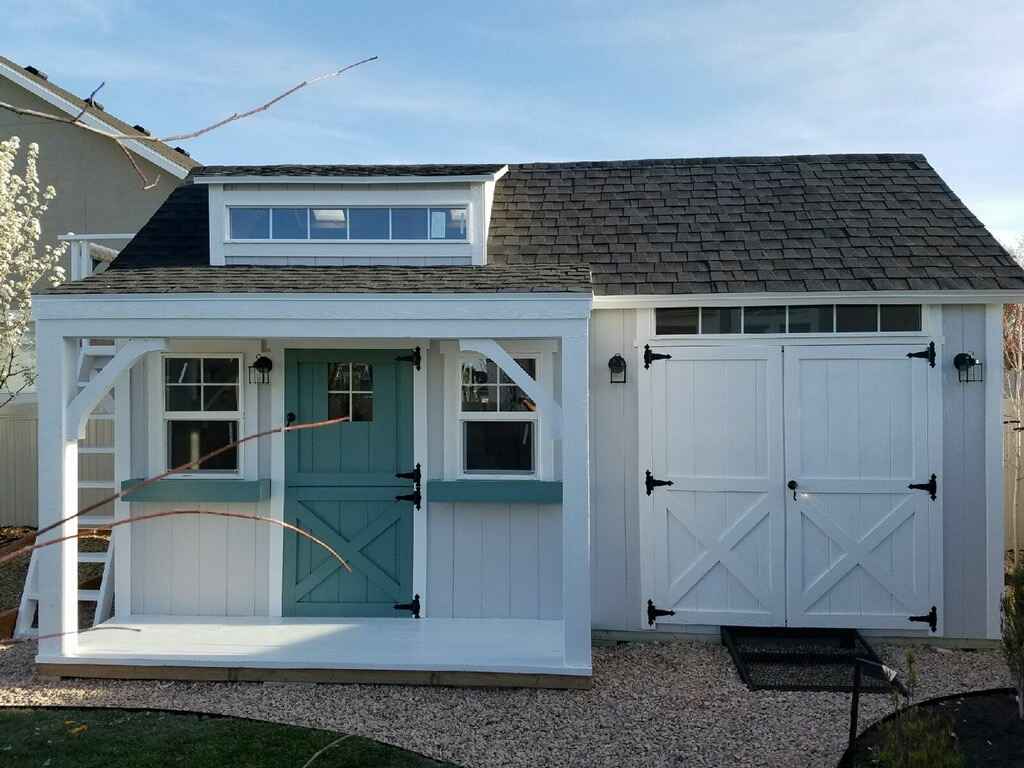 White playhouse shed with porch, teal Dutch door, transom windows, and double barn-style storage doors on a gravel pad