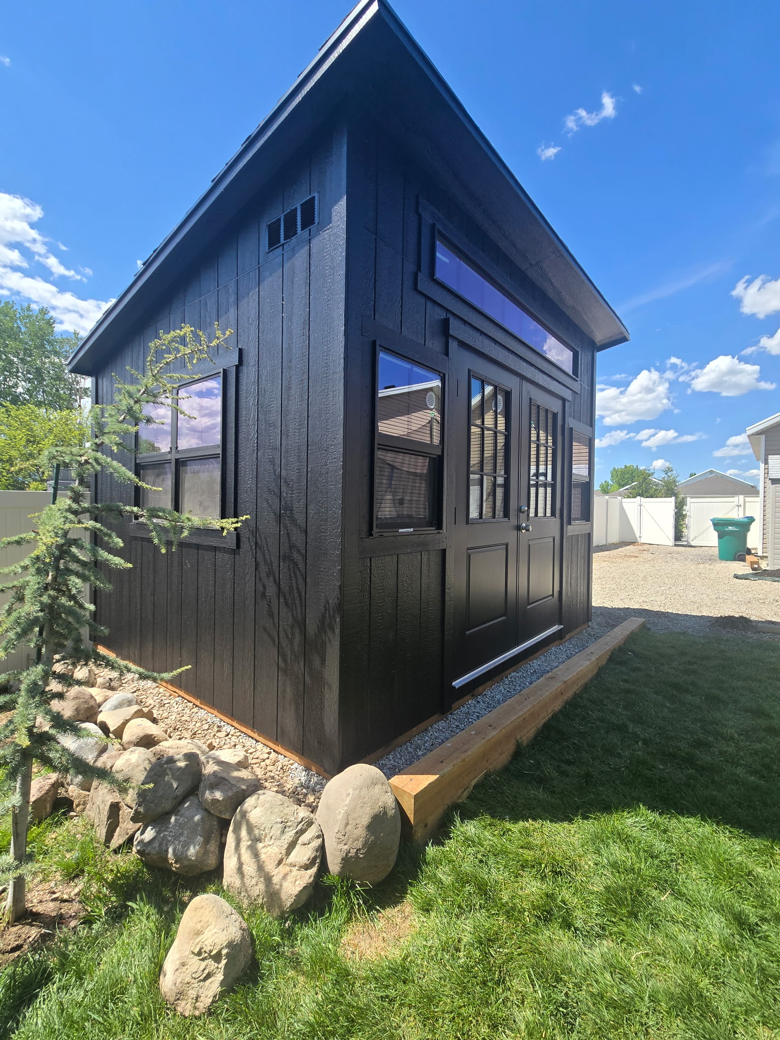 Modern black 10×12 lean-to shed with transom windows and double doors in a landscaped yard, built for custom storage solutions