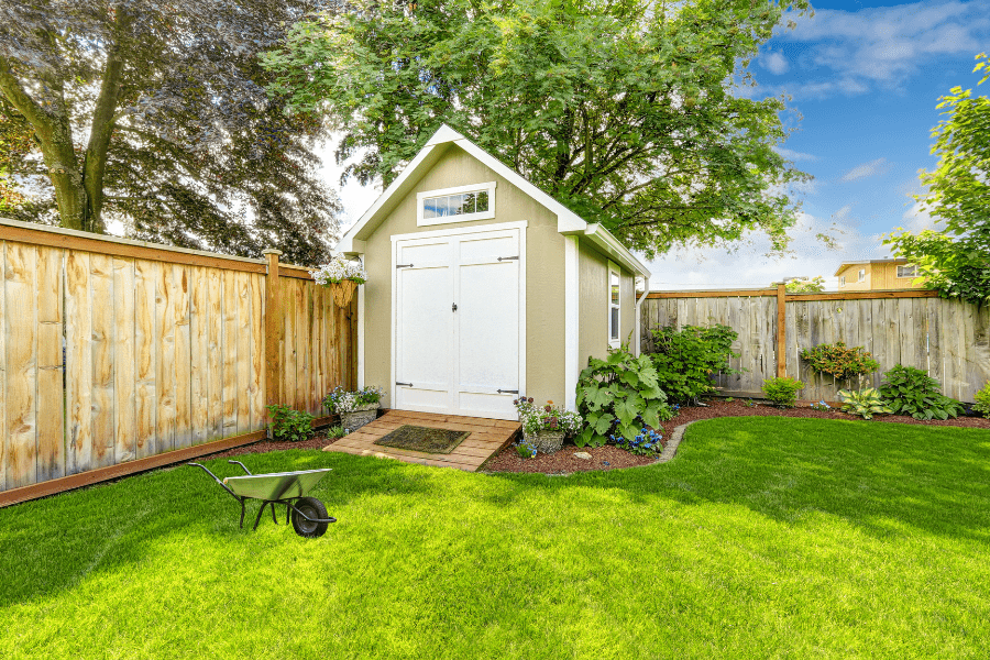 Light green garden shed with white doors and brick paver ramp in a landscaped backyard. Focus keyword: shed with ramp