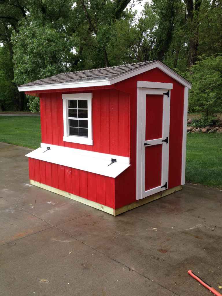 Red custom chicken coop with white trim, shingled roof, window, nesting-box lids, and door, set on a concrete pad