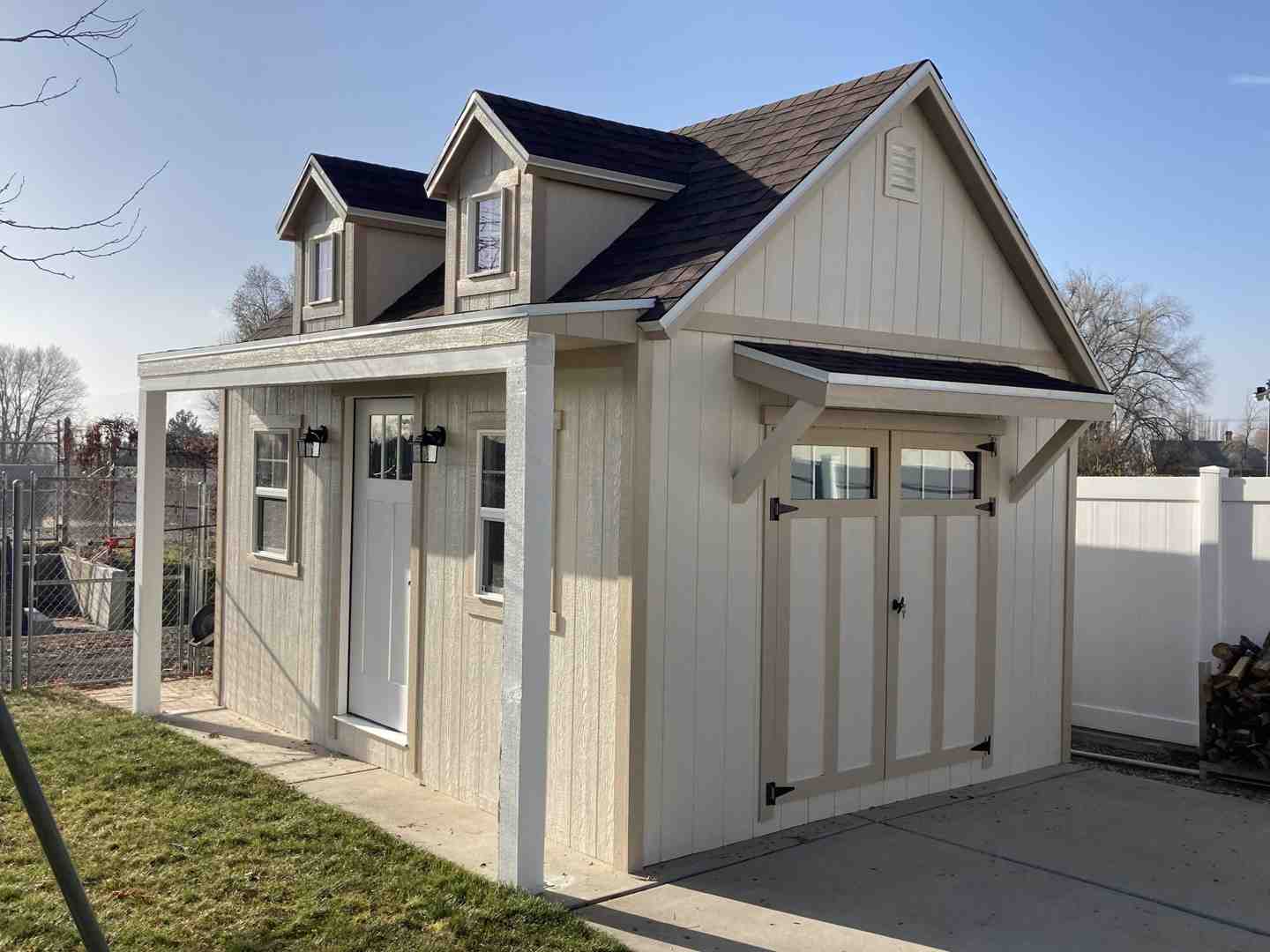 Beige custom storage shed featuring dormer windows, double doors, and a side porch with lighting