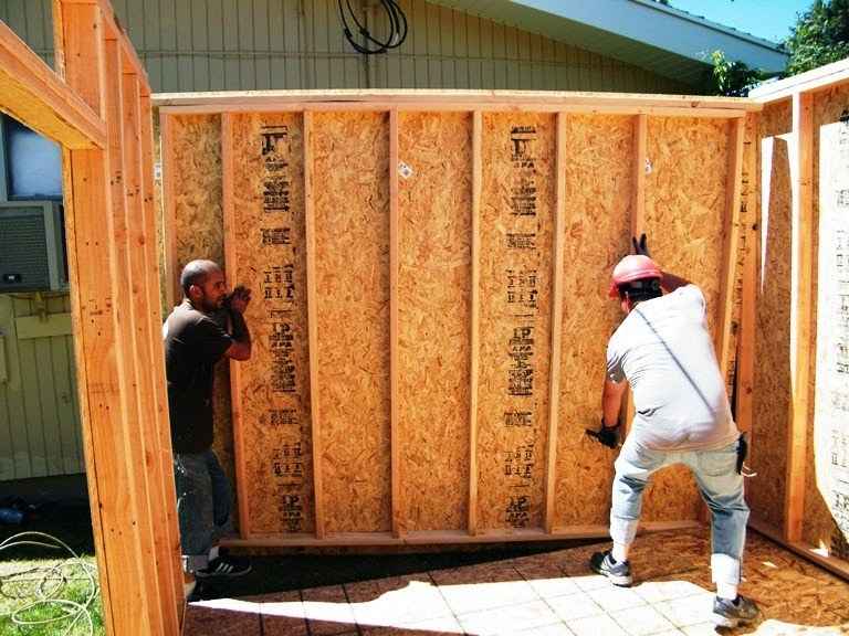 Two builders lift a framed OSB wall panel into place for a backyard shed, aligning studs on the slab before fastening and bracing.