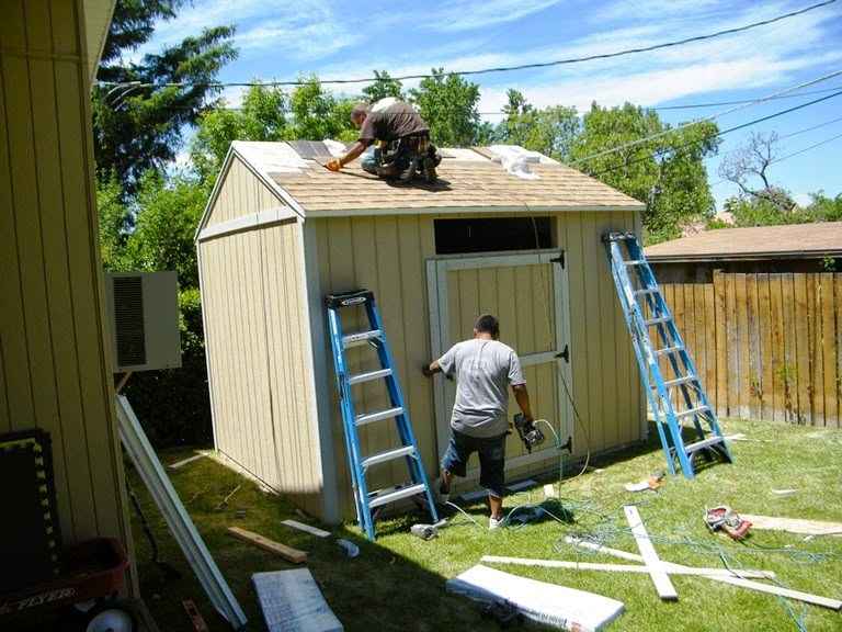 Two men installing shingles and exterior trim on a wooden shed, showing final steps in how to build a small storage shed