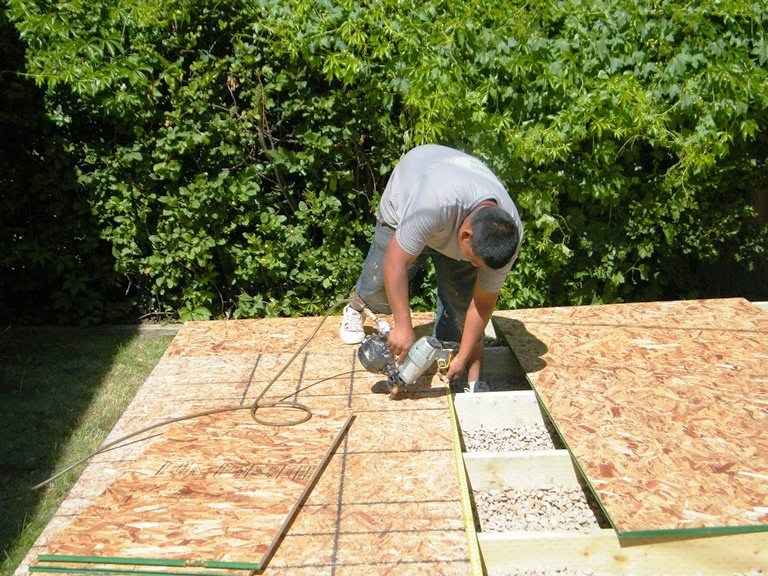 Worker cuts OSB sheathing for a DIY shed floor; joist bays filled with gravel, leveled base below, circular saw and power cord visible.