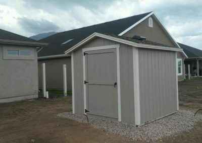 Small gray backyard shed with white trim on gravel beside a home, representing affordable local rent to own sheds options.