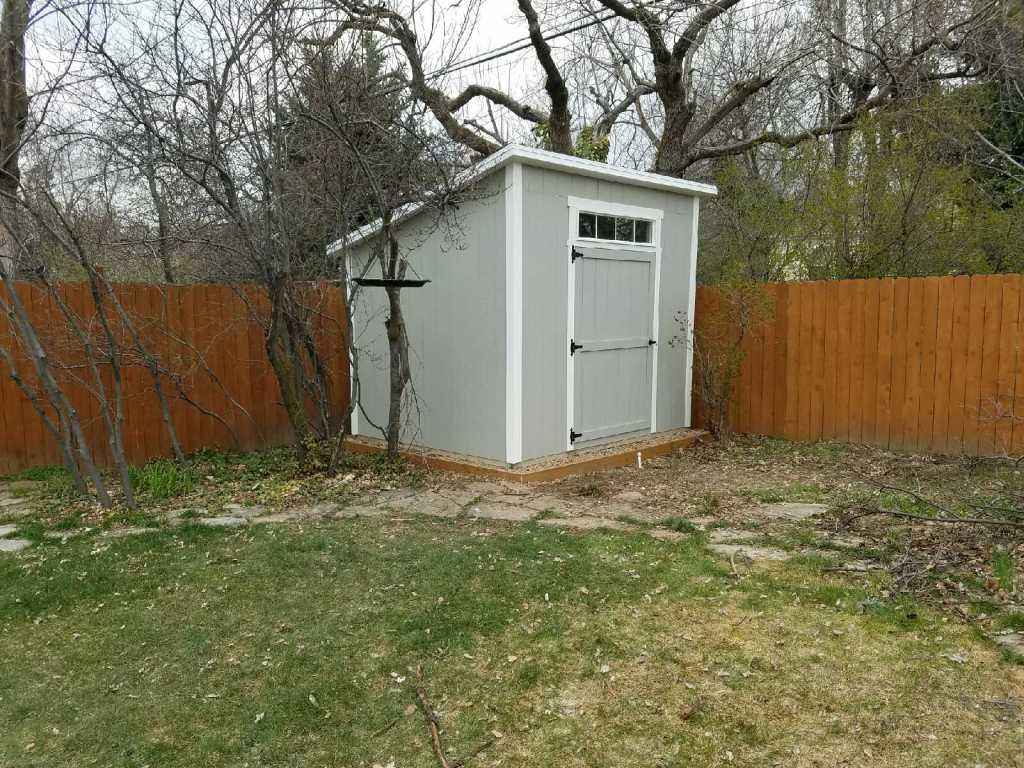 Backyard 8x8 lean to shed with gray siding, single door and transom window, on wood base beside cedar fence and trees.