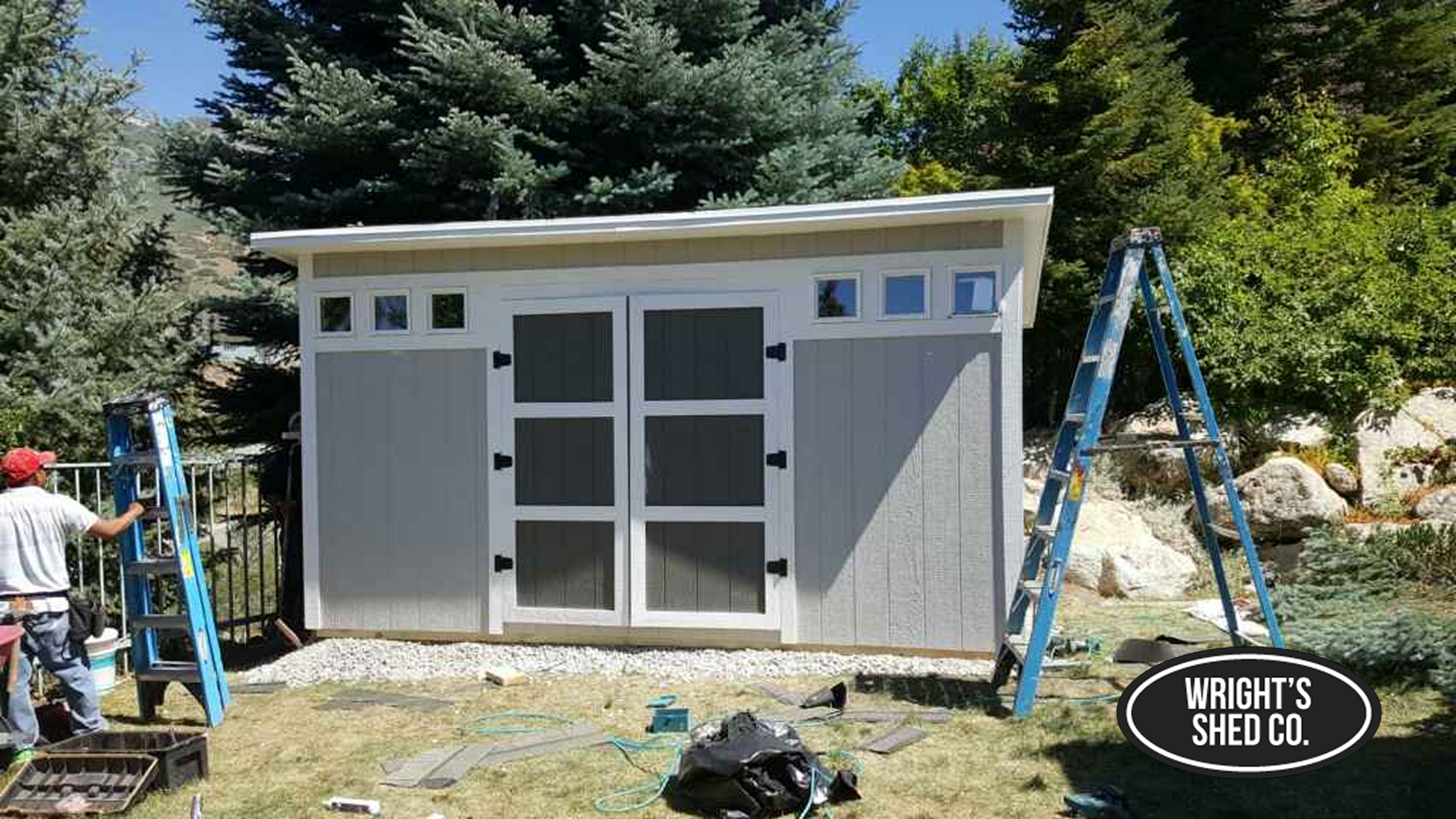 Wood Shed Installation in Utah Workers installing a wood shed in a Utah backyard, showing ladders, tools, and the completed shed exterior with Wright’s Shed Co. branding.