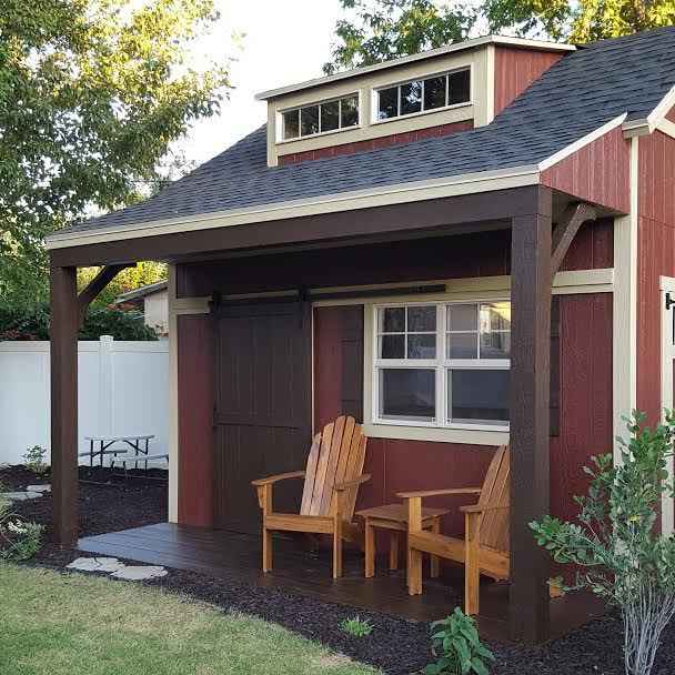Red she shed with covered porch, barn-style sliding door, dormer windows, and two Adirondack chairs on a decked patio