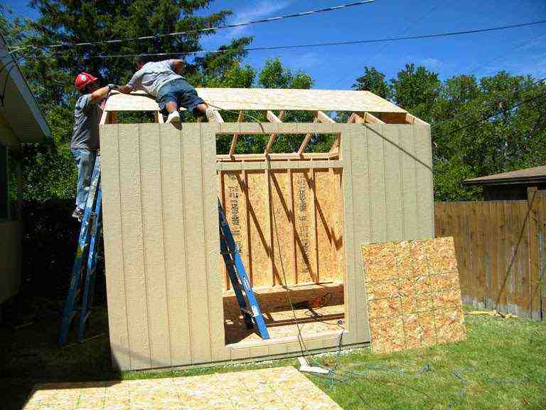 Two workers attaching OSB roof panels to a pre-cut shed kit frame using ladders