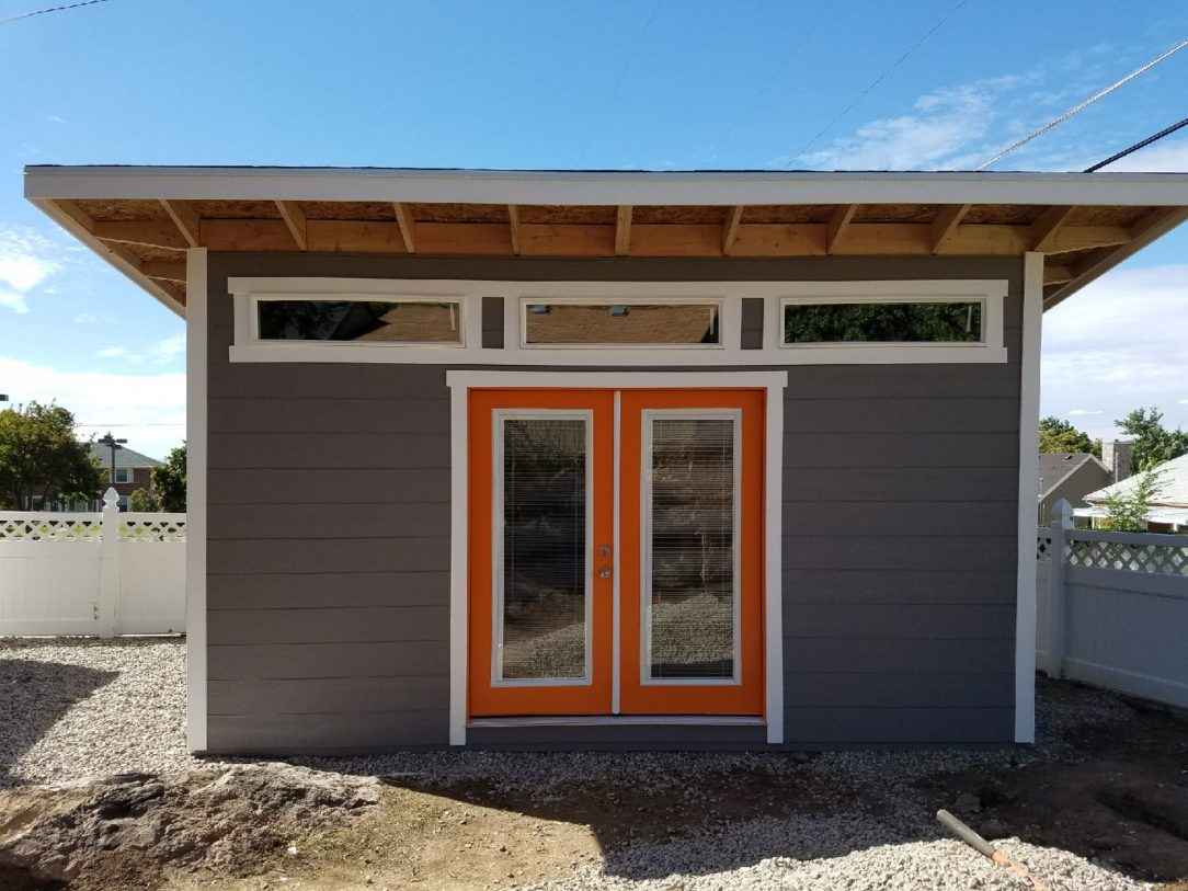 Modern gray lean-to shed featuring exposed overhangs and bright orange double doors with transom windows for a stylish backyard look.