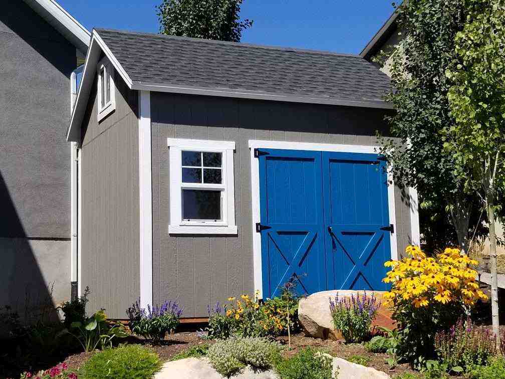 Gray backyard storage building with bright blue double doors and flower garden in front