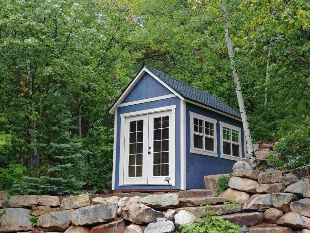 Blue orchard shed with white trim, double patio doors, and large windows, set on a stone terrace surrounded by trees