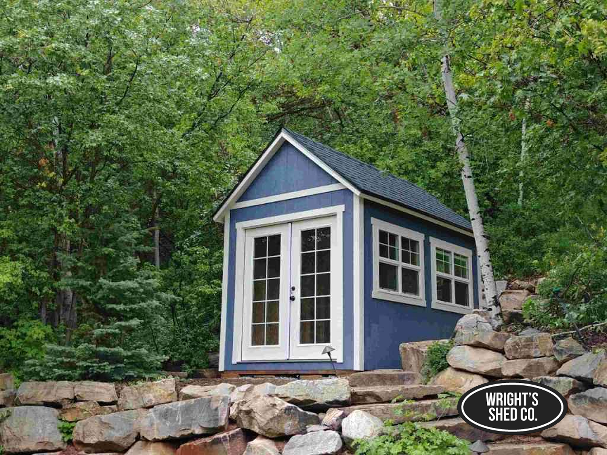 Elegant Wooden Mini Shed in a Forest Setting Blue wooden mini shed with white trim and double doors nestled among trees on a rocky landscaped slope, built by Wright’s Shed Co.