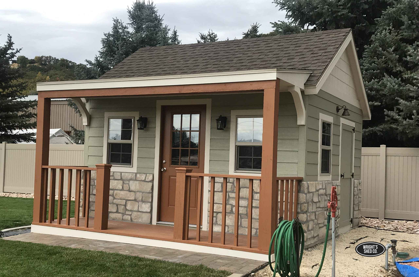 Cozy She Shed with Porch in Utah She shed with a front porch, railings, stone accents, and wood trim in a landscaped Utah backyard, built by Wright’s Shed Co.
