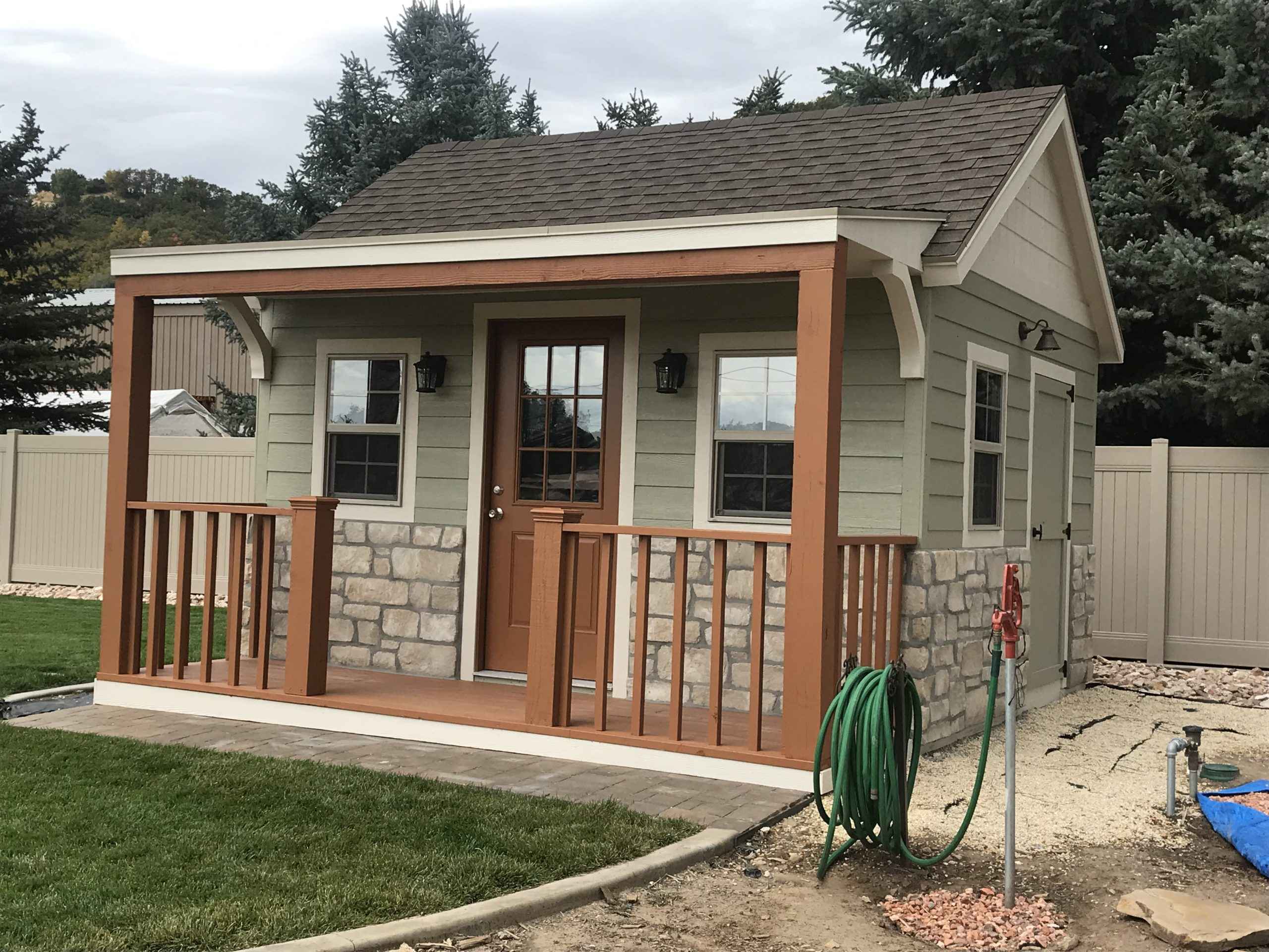 Beautiful Backyard Shed With Porch and Railings A stylish backyard shed with a front porch, railings, stone accents, and warm wood trim, perfect for homeowners seeking shed with porch ideas.