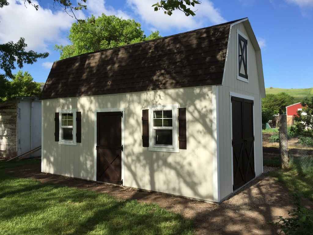 Sunny view of a cream farm shed with brown gambrel roof, double doors and shuttered windows, on grass beneath trees