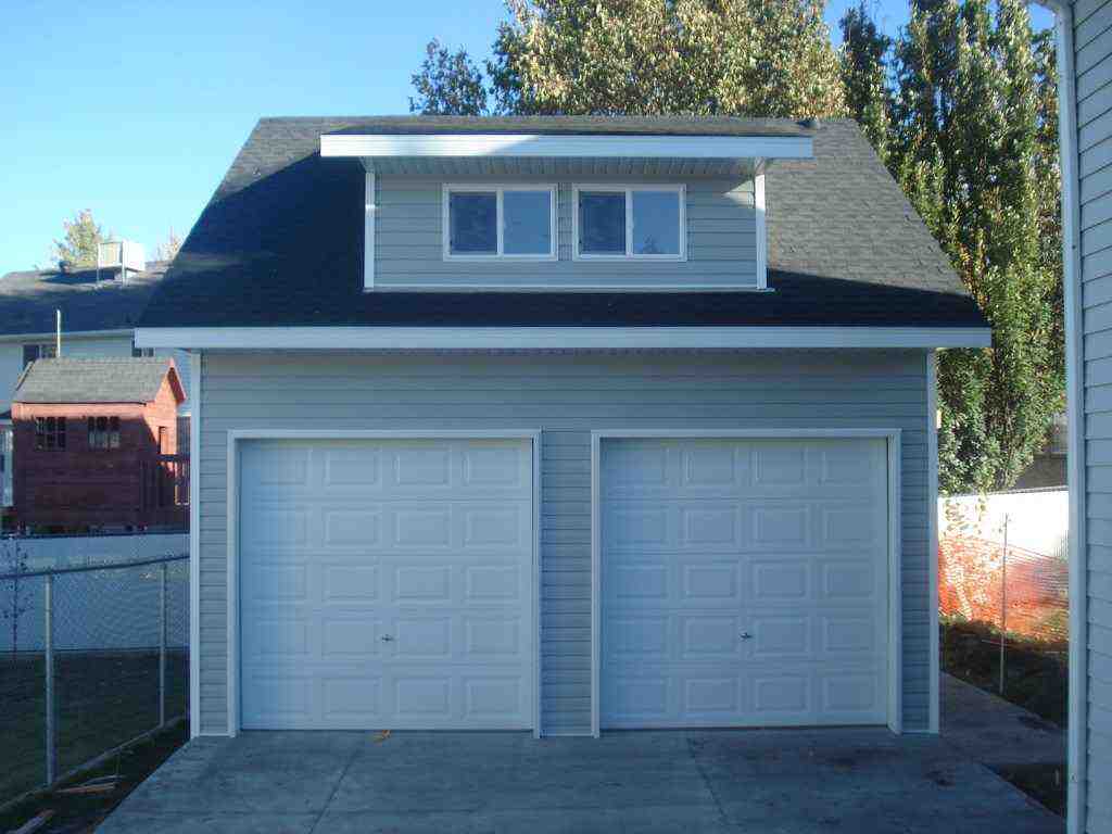 Detached two-car dormer garage with light gray vinyl siding and white roll-up doors