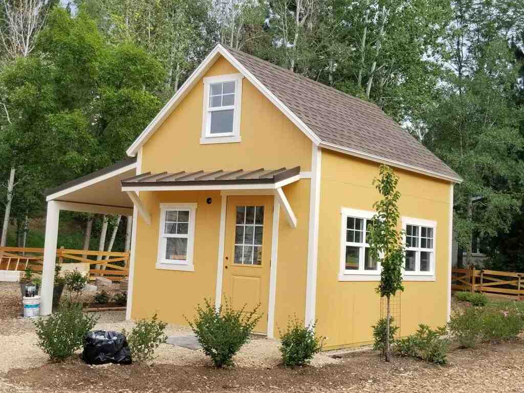 Bright yellow custom storage shed with front porch, side awning, and white trim set in a landscaped yard