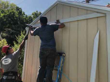 Two builders installing white trim on the gable end of a beige wooden shed during the finishing stage of a Wright’s Shed Co. DIY shed kit assembly.