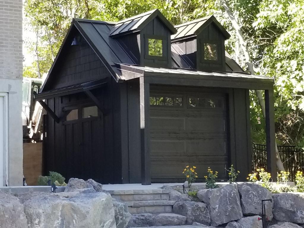 Modern black shed with dormer windows and garage door beside stone steps and trees, built for custom storage solutions