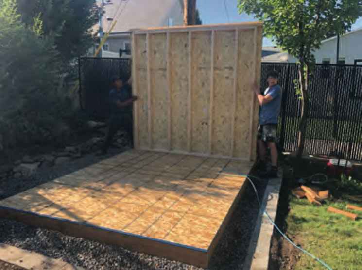 Installing the Walls on a Shed Kit Two people lifting and positioning a framed wall section onto a shed floor, demonstrating a key step in how to assemble a shed kit properly.