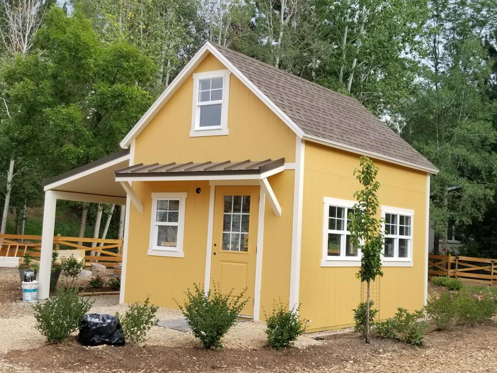 Bright yellow custom storage shed with white trim and covered side porch, built for versatile use in a landscaped outdoor space. 