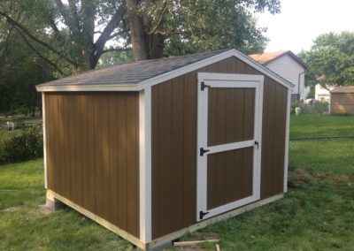 Brown wooden DIY shed kits with white trim, double doors, and gable roof installed on a grassy backyard lot.