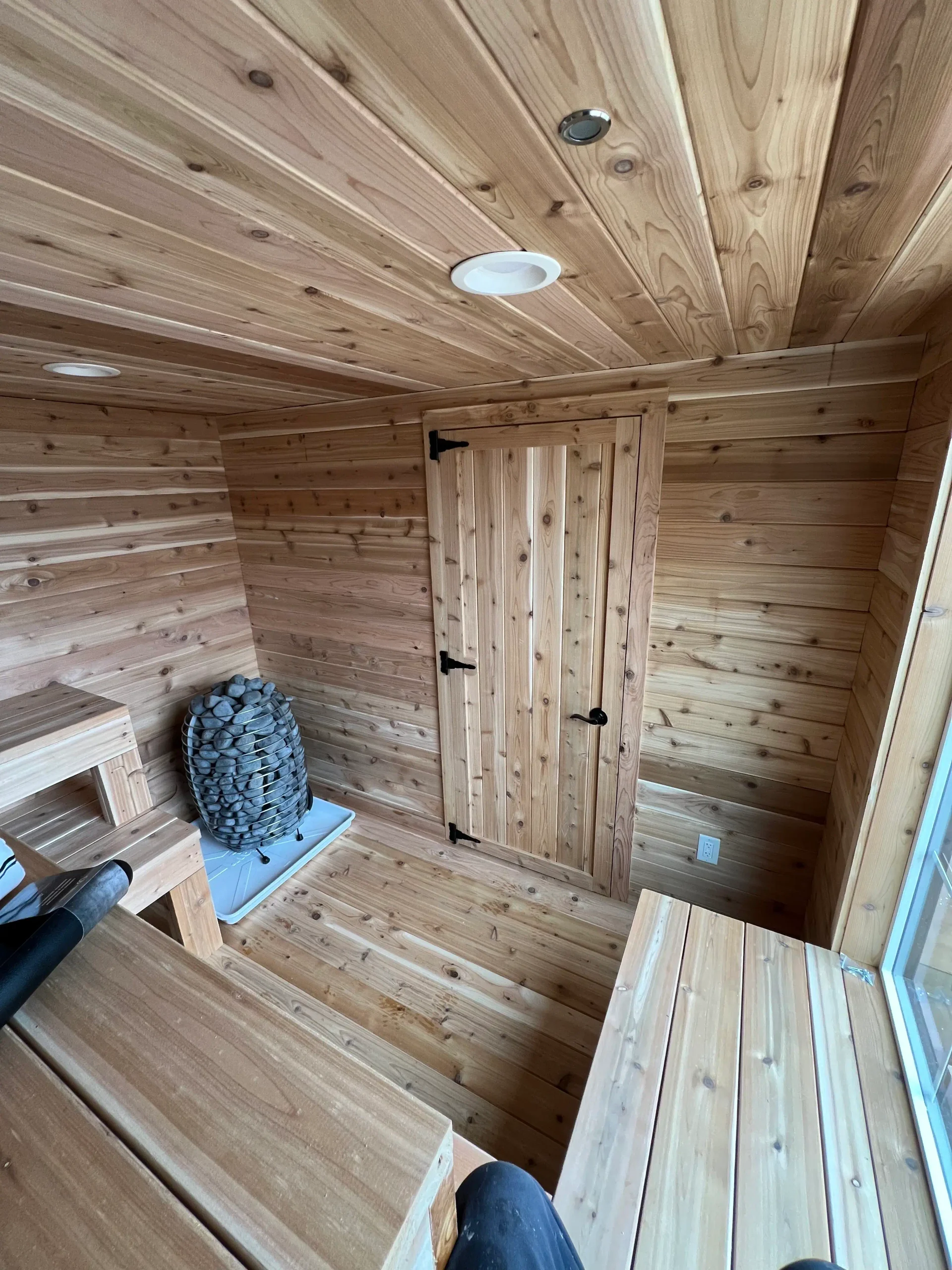 Cedar sauna interior with wood walls, ceiling, benches, stone heater, and natural wood door beside a window.