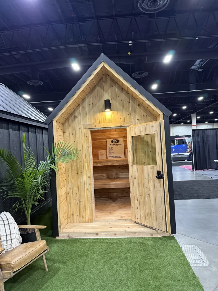 A finished wood sauna display shows how a custom sauna is built with cedar walls, benches, a door, and metal roof.
