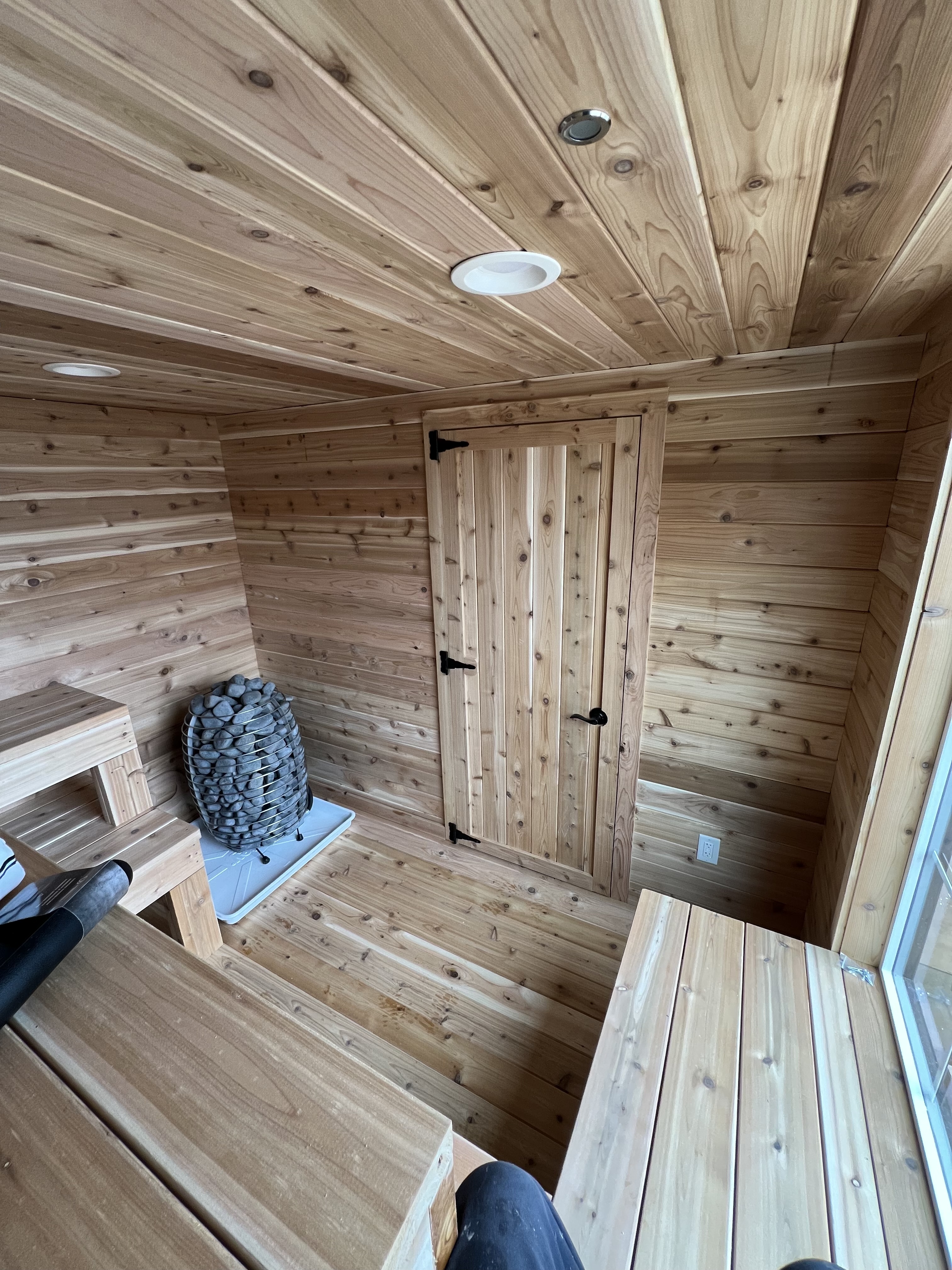 Interior view of a custom cedar sauna by Wright's Shed Co. with cedar tongue-and-groove walls and ceiling, recessed lighting, a cedar door with iron hardware, two-tier bench seating, and a stone heater on the floor.