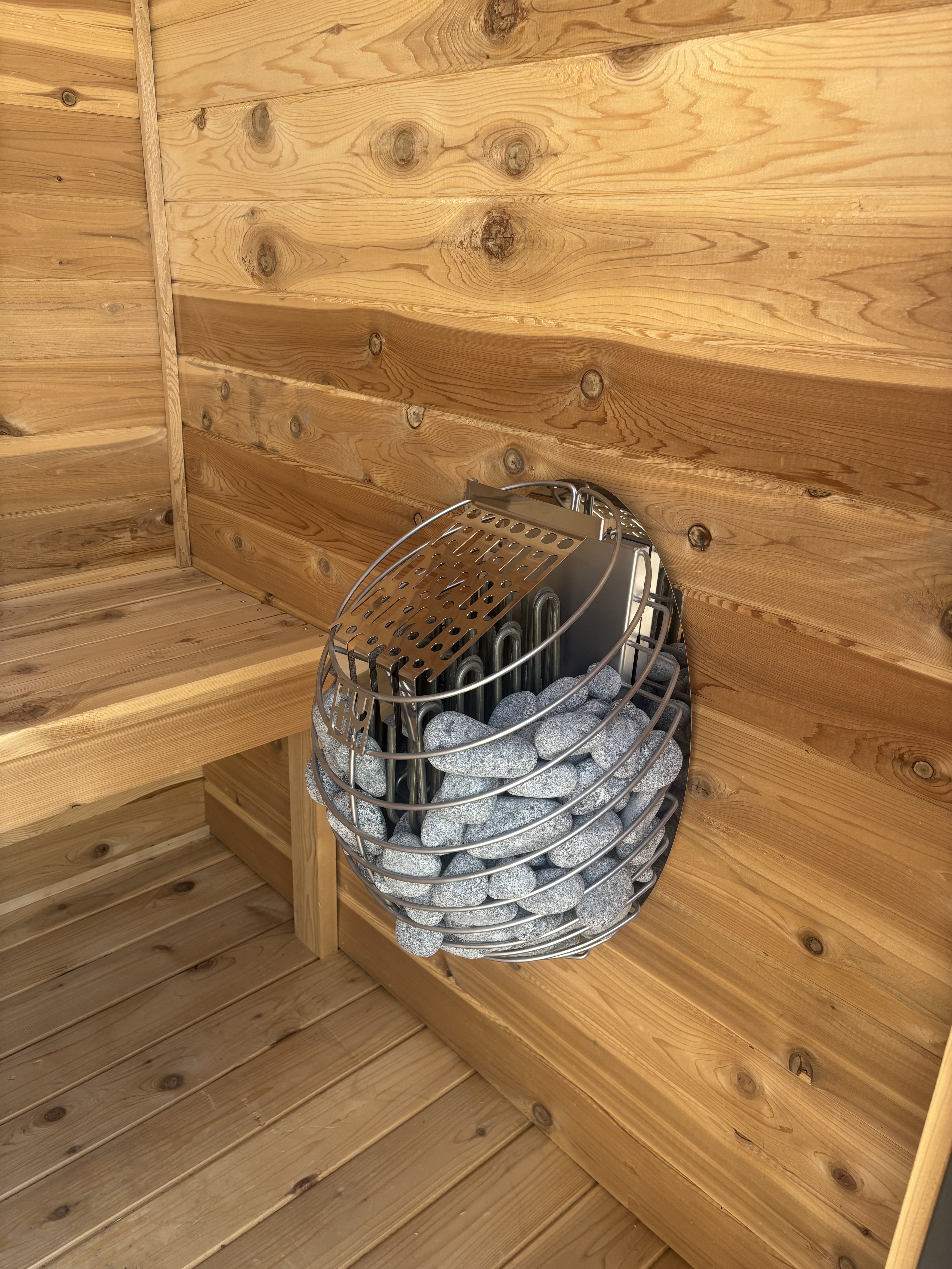 Close-up of a stainless basket heater filled with sauna stones inside a Wright's Shed Co. cedar sauna, showing the natural grain and knots of the cedar plank walls.
