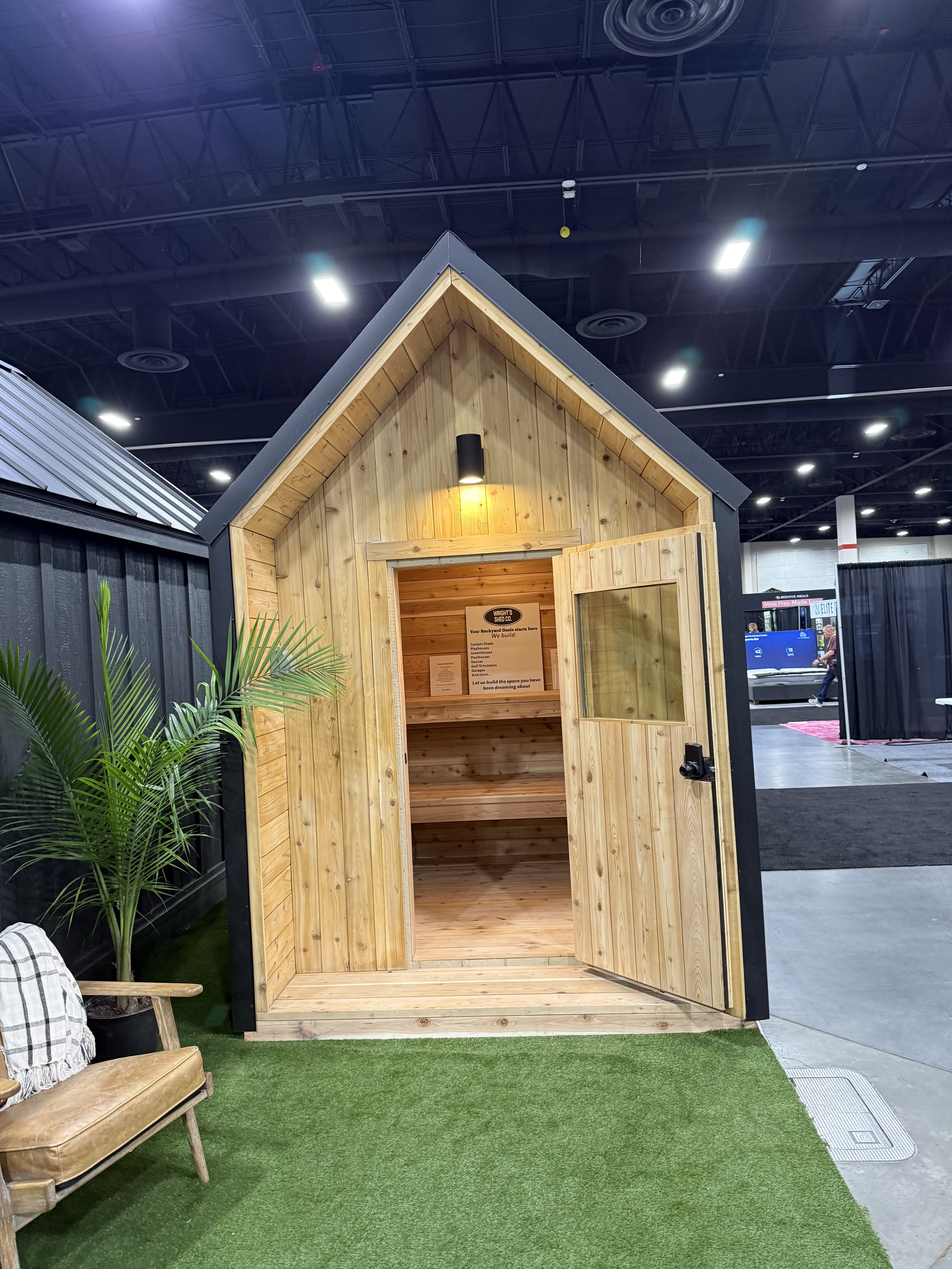 Custom cedar backyard sauna by Wright's Shed Co. with peaked A-frame design, black metal trim, and an open door showing the tongue-and-groove cedar interior with bench seating.
