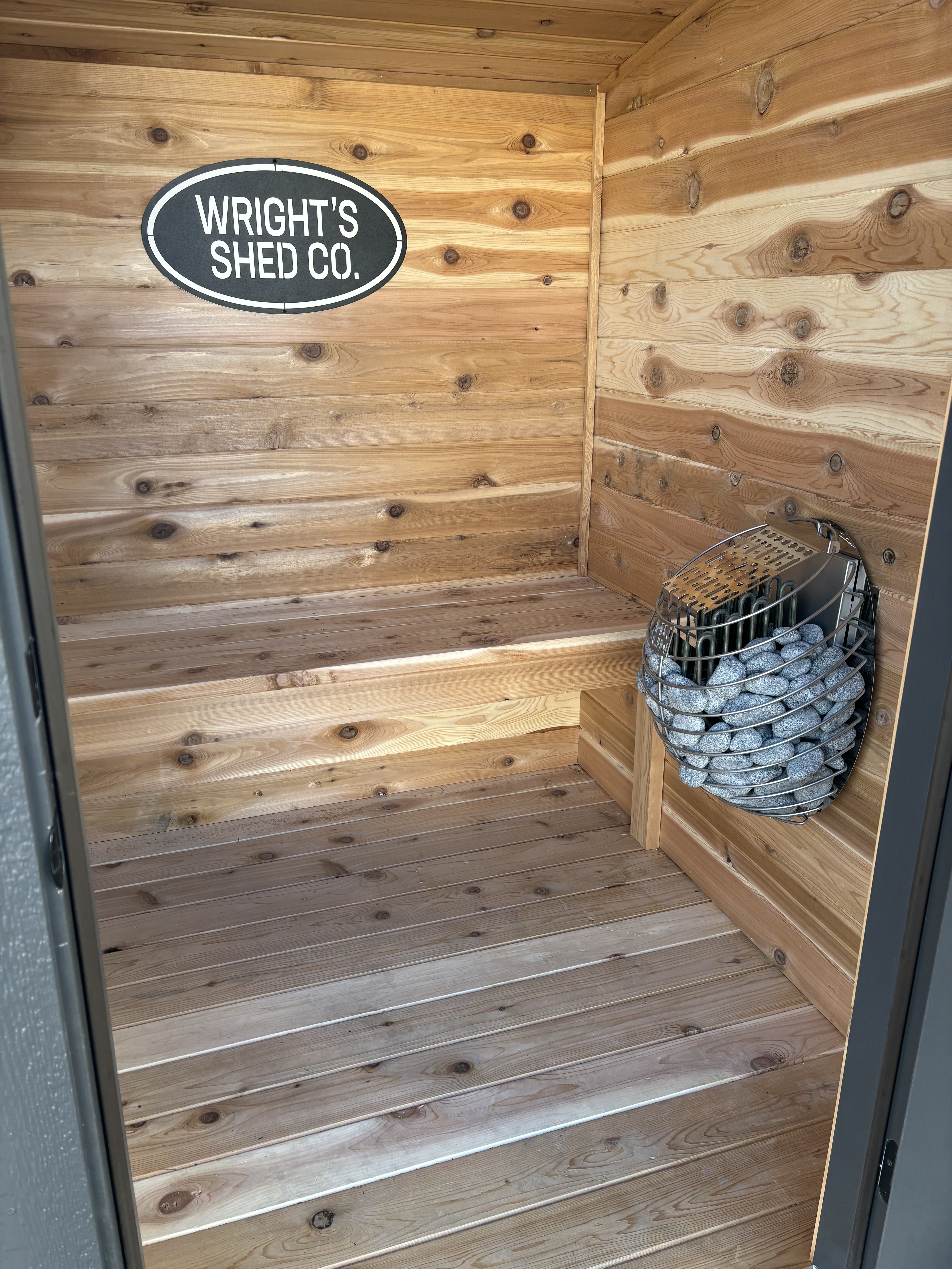 Inside a custom cedar sauna built by Wright's Shed Co. in Utah, showing tongue-and-groove cedar walls, L-shaped bench seating, and a wall-mounted heater with sauna stones.
