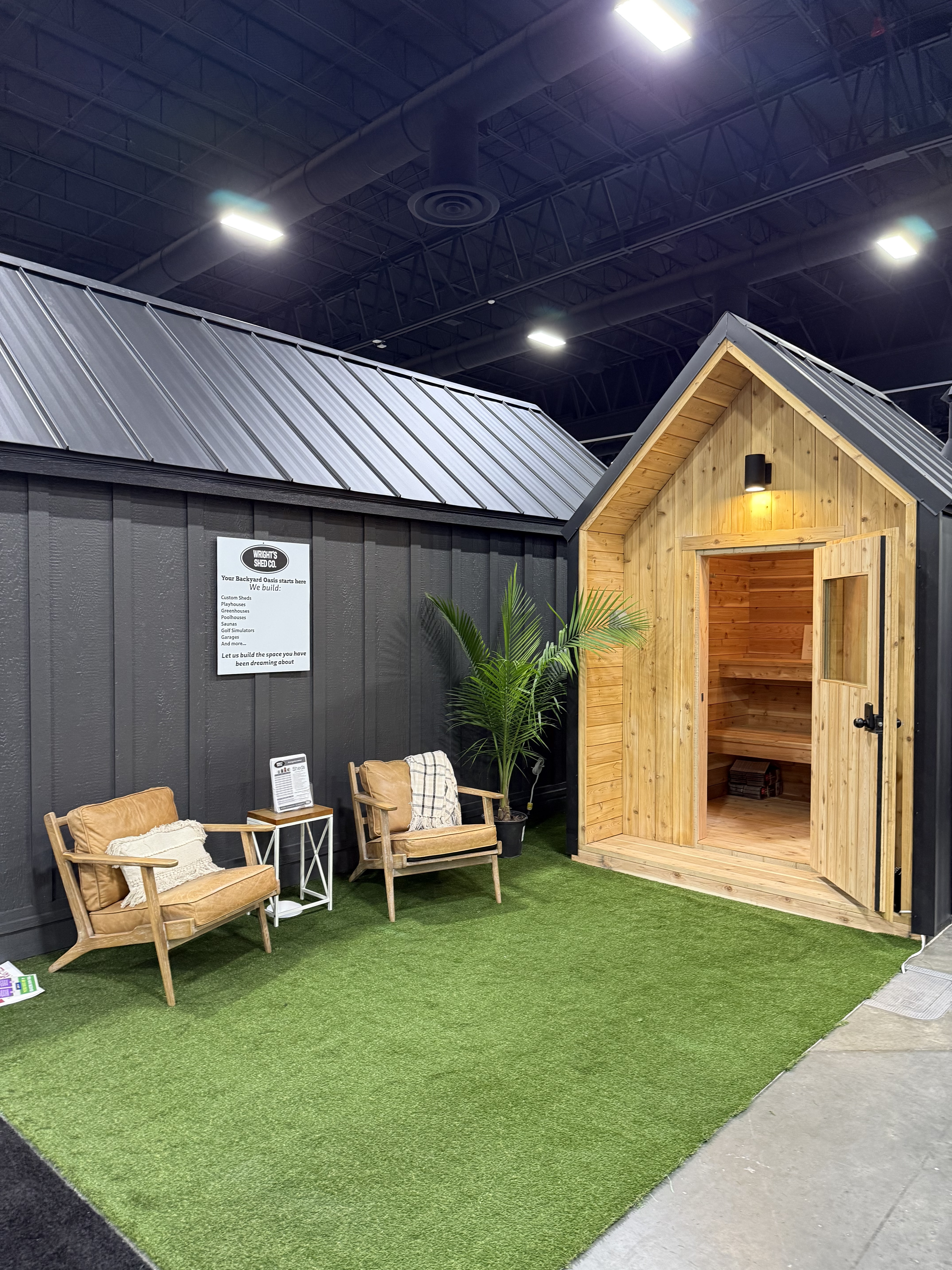 Wright's Shed Co. home show display in Utah featuring a custom cedar backyard sauna with the door open, alongside a dark board-and-batten outbuilding, leather lounge chairs, and signage.